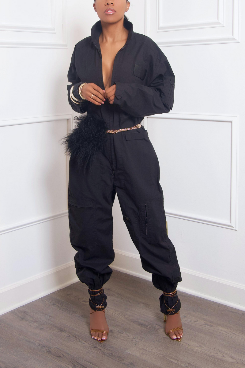 A woman in a black jumpsuit with gold trim and strappy heels, holding a Mongolian fur purse, posing against a white paneled wall.
