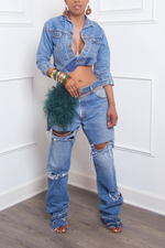 A woman wearing an upcycled Denim Asymmetrical Jean Jacket, ripped jeans with patchwork details, and accessorized with bangles, earrings, and heeled sandals stands against a white background